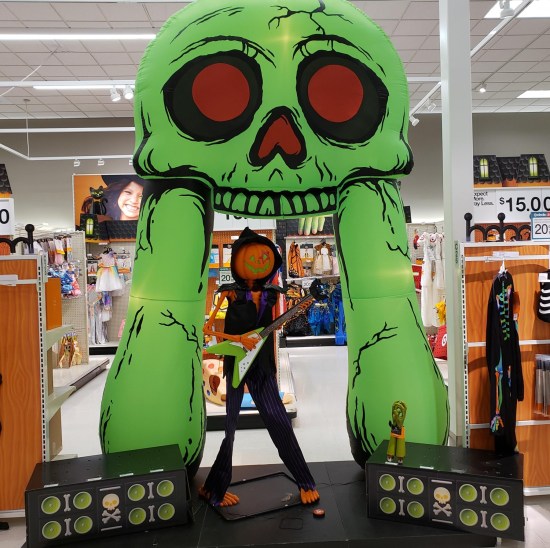 Department store display: orange skeleton with pumpkin head in black rock-star clothing, playing guitar beneath what looks like a neon green mammoth skull with red eyes and nasal cavity.