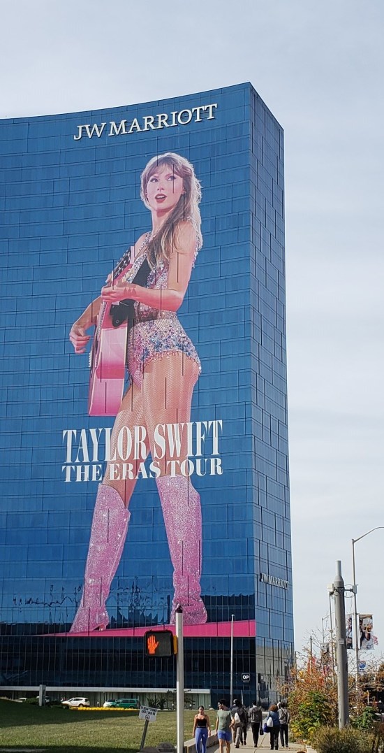 Giant Taylor Swift mural on the front of a Marriott whose face is all blue glass. She has a pink guitar.