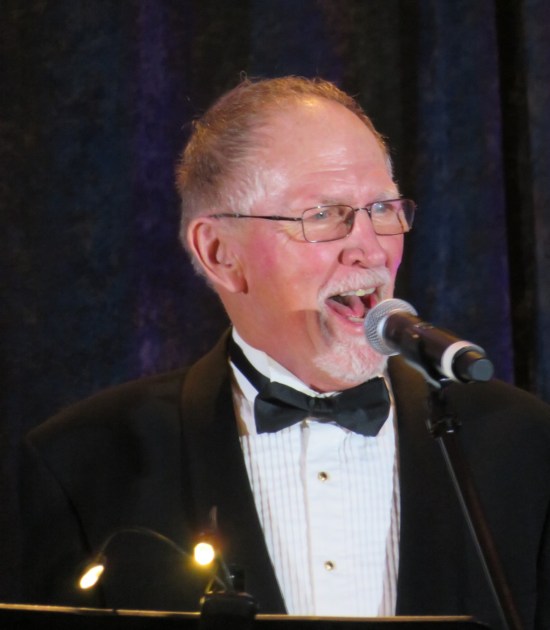 Vaughn Armstrong sings, wearing glasses and a tux.
