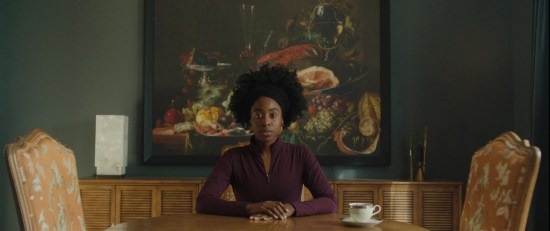 Kirby sitting alone at one end of a fancy table in front of a large, banal still-life fruit painting. She stares at unseen people on the opposite end, an untouched teacup beside her.