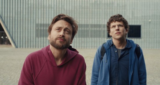 Two thirtysomething Jewish men staring at offscreen WWII remembrance statues in solemnity.