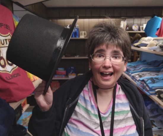 Anne in a gift shop with dark brown wood-paneled walls, smiling and waving a top hat.