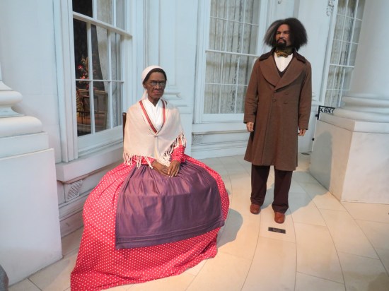 Porch of the faux White House with an older Black lady sitting in a red skirt and off-white shawl. Standing next to her, 