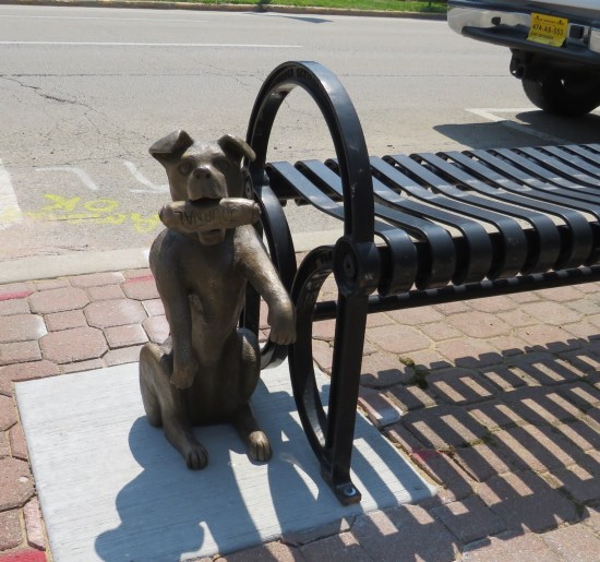 Statue of a doggie sitting up next to a real bench with a rolled-up Journal newspaper in its mouth.