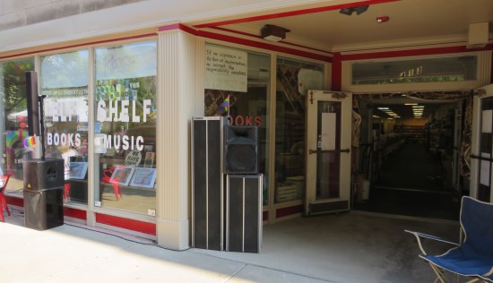 Downtown bookstore with both doors propped open, lots of shelves visible through the doorway, and large speakers standing out front.