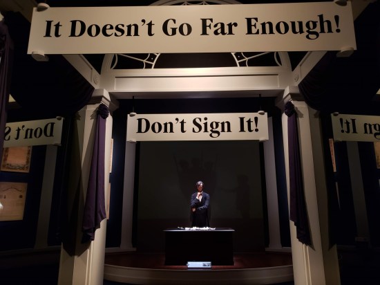 Distant statue of Lincoln at a desk thinking. Above the path leading to him are signs such as "It doesn't go far enough!" and "Don't sign it!"