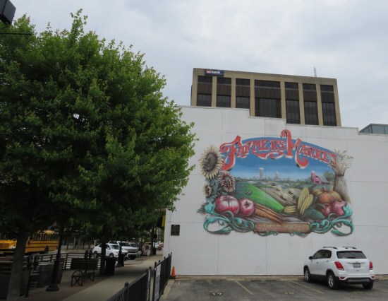 Parking lot next to a white building with a farmers' market mural, labeled "Established 1999". The adjacent street is lined with healthy trees.