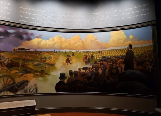 Curved mural of Lincoln giving the Gettysburg Address at a cemetery. His handwriting is blown up and adorns the wall above.