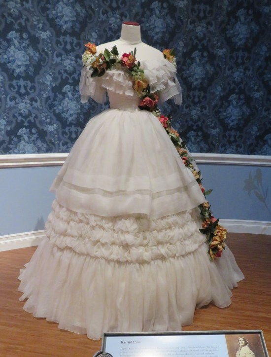 All-white dress with wide neckline off the shoulders and a long, singular string of flowers twined around the dress from bosom to a foot from the floor.