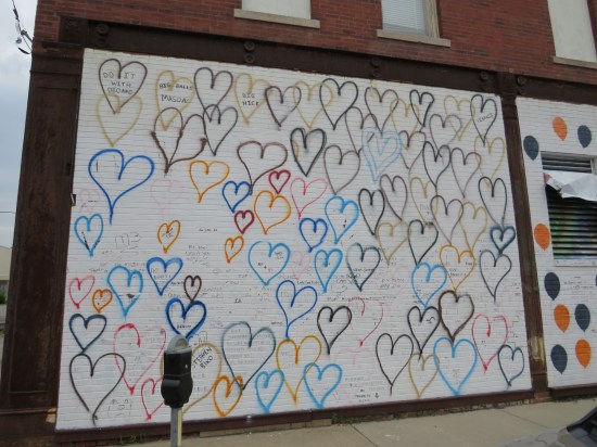 White wall with hand-drawn heart outlines in different colors. Messages written in them include "Stephen Biko", Do It With Dionne", "Big Balls Mason", and "Do What U Love".