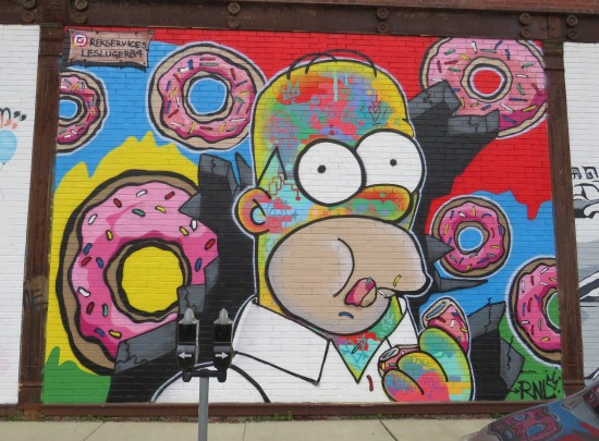 Brick wall mural of Homer Simpson eating one of many pink-frosted donuts raining upon him from above. Psychedelic tattoos cover his open yellow flesh.