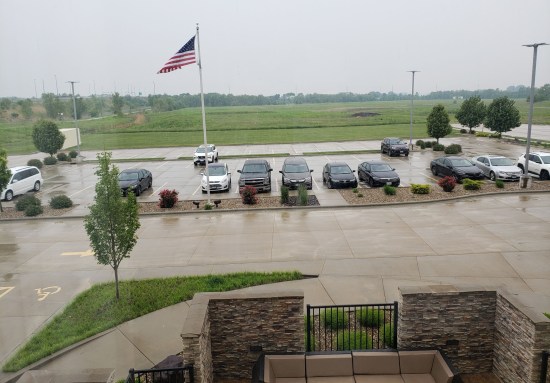 A rainy parking lot with a large, well-kept grassy field beyond. Near the camera is the edge of a walled courtyard with a cushioned patio couch.