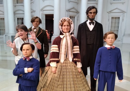 Statues of the Lincoln family (Abraham, Mary and their three sons) in front of an indoor replica of the White House facade. Anne stands between two of the boys, doing jazz hands.