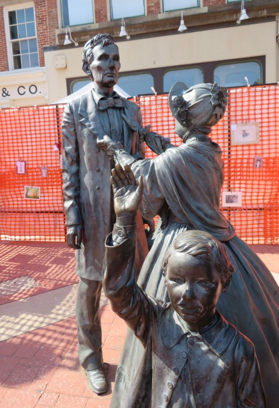 Statues of Lincoln, Mary, and young Robert getting ready for church. Around them is a temporary wall of orange mesh with children's drawings hung on it.