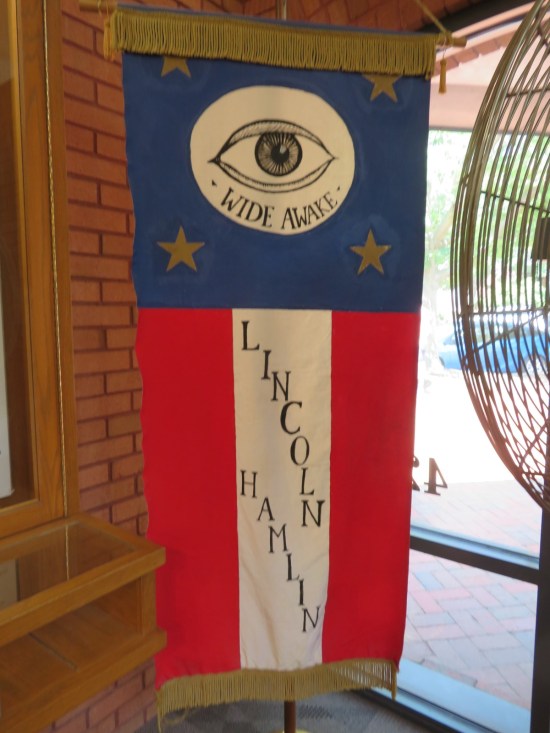Vertical banner with a blue field, four gold stars, and one white stripe with Lincoln's and Hamlin's names on it, between two red stripes. On the blue field is a white circle with an eyeball and the slogan "WIDE AWAKE".