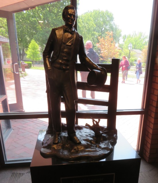 Indoor bronze statue of Lincoln nicely dressed, top hat in hand, leaning against a fence.
