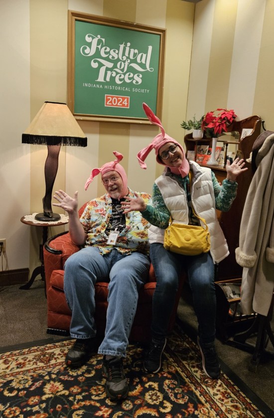 Two people sitting in the old man's armchair wearing pink bunny hats next to the leg lamp. A Festival of Trees sign hangs on the wall.