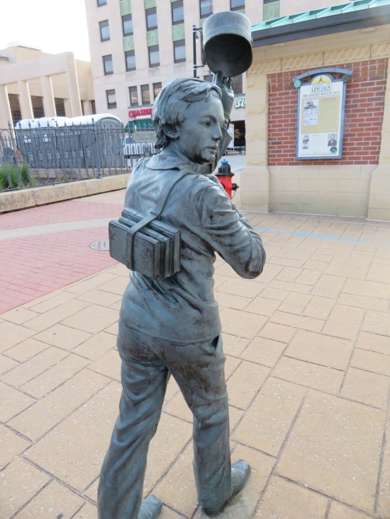 Statue of a schoolboy toting books on his back with a strap and doffing his old-timey cap. In the distance is a Cold Stone Creamery.