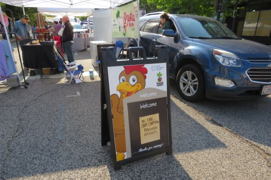Sandwich board advertising the Old Capitol Farmers' Market with a rooster drawn on it. Behind is one of its tents; an SUV drives by with a kid hanging out the window.