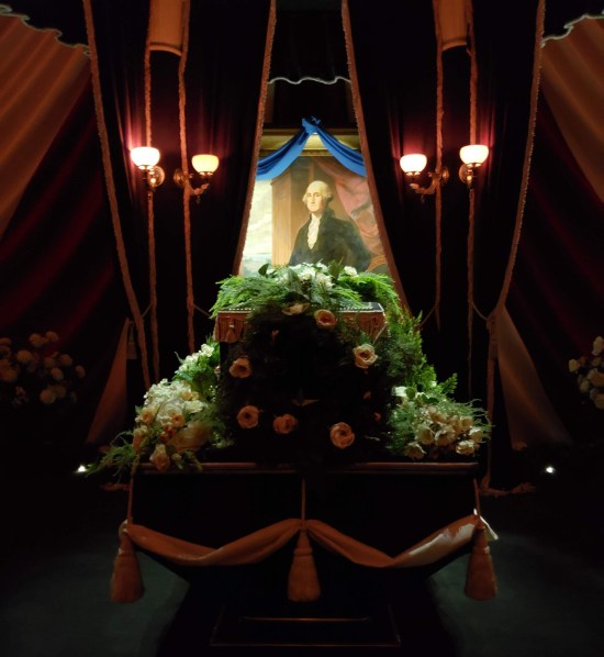Front of Lincoln's shrouded coffin with a wreath and white flowers. Behind it hangs a George Washington painting.