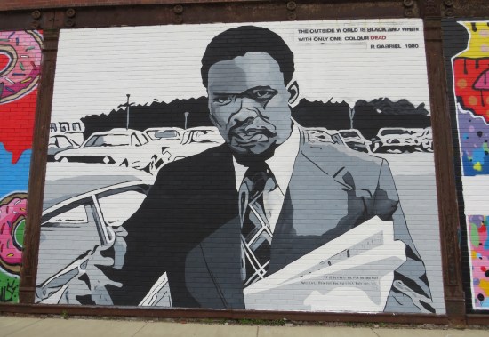 Black-and-white mural of Steve Biko standing next to a car with a file folder in his arm. Peter Gabriel lyric at the top: "The outside world is black and white with only one colour dead."