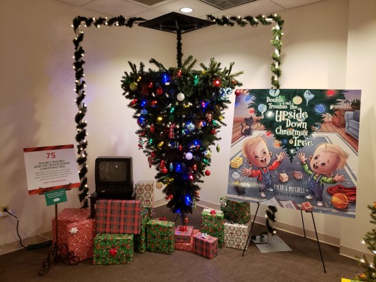 Upside-down Christmas tree with presents under it, a TV turned off, a blown-up children's book cover, and a rectangular evergreen border with lights around the whole display. 