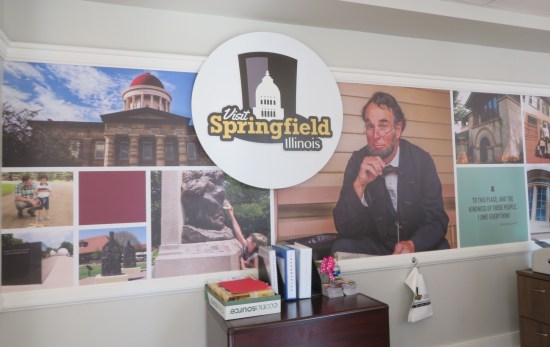 Visitor center with Lincoln photos on the wall -- cosplayer, Old State Capitol, bust, "Acts of Intolerance" installation. Center circle says "Visit Springfield, Illinois". Farther down, a Lincoln quote: "To this place, and the kindness of these people, I owe everything."