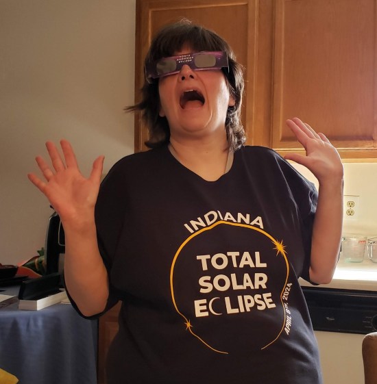 Anne in our kitchen wearing eclipse glasses and an Indiana Total Solar Eclipse T-shirt, doing jazz hands.