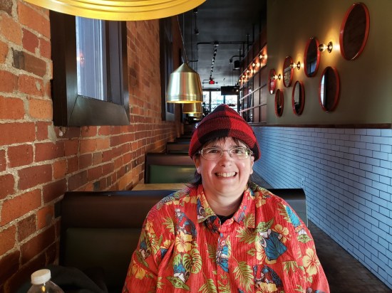 Anne sitting in a sandwich shop in a red-and-black flannel cap and a Mandalorian tropical shirt. She's smiling really big.