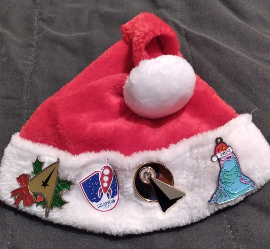 Santa hat with four pins: GalaxyCon 2024, a Starfleet symbol with mistletoe and holly, a triangle intersecting with a circle that I don't get, and Murf from "Star Trek: Prodigy" wearing a 2022 Santa hat.