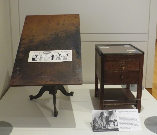Old wooden drawing board and side table in a museum exhibit. A Dick Tracy strip is mounted in the middle of the table.