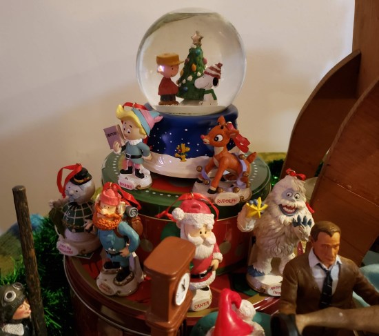 A small and a large Christmas cookie tin stacked in a tiny ziggurat on a festive table. On top is a Peanuts snow globe. On each level stand ornaments of the cast from Rankin-Bass' "Rudolph the Red-Nosed Reindeer".