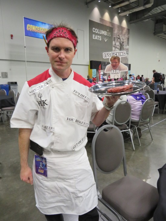 costime: trainee chef with red bandanna, white smock covered in Gordon Ramsay quotes, profanities censored. He holds a serving tray with a fake steak and a picture of Ramsay yelling "IT'S F-ING RAW!"