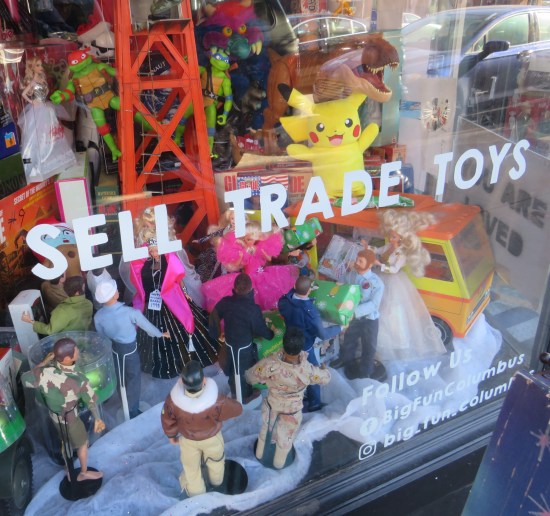 Store window display: Pikachu doll stands on a toy van surrounded by several 12-inch dolls facing toward him, away from us. Window lettering reads "Sell Trade Toys".