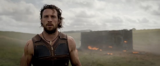 Leather-clad long-haired hunting guy stands on an African plain, slack-jawed. Behind him, an overturned truck burns.