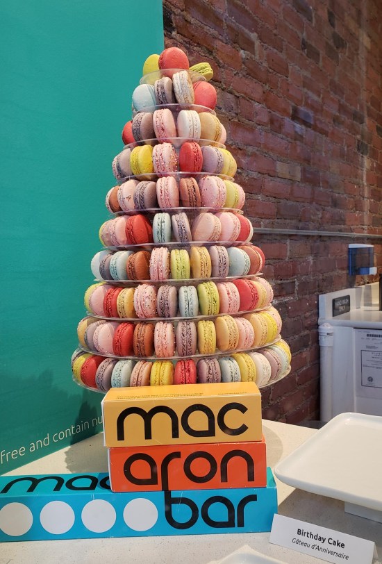 Dozens of macarons in a pyramid on a plate. Sign in front of display for Macaron Bar, teal walls behind it.