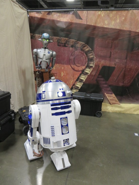 R2-D2 and a weird silver angular droid far behind him. The background is a large photo of a Jawa Sandcrawler.