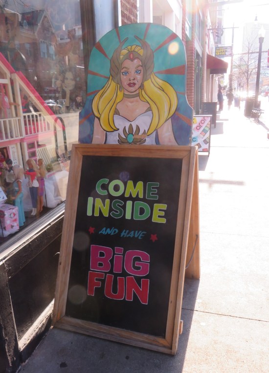 A-frame chalkboard next to shop reading "Come inside and have big fun." Drawing of She-Ra on top.