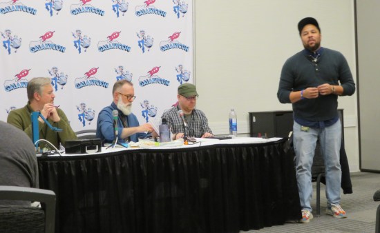 Three white guys at a panel-room table and one Black guy off to the side, all of them gentlemen.