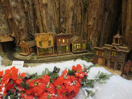 Intricate wood model of mountainside with five Old West establishments and train tracks. Poinsettias are placed at the table front.