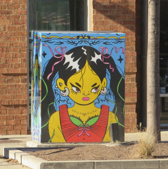 Electrical box painted with a yellow-skinned young Hispanic woman in a sleeveless red top and decorations floating around her.