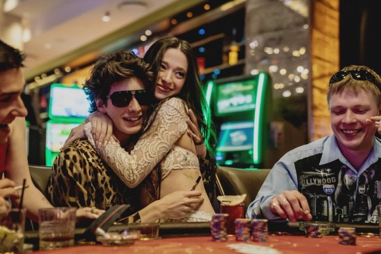 A smiling young couple sit at a Vegas card table, she in his lap, arms around his neck.
