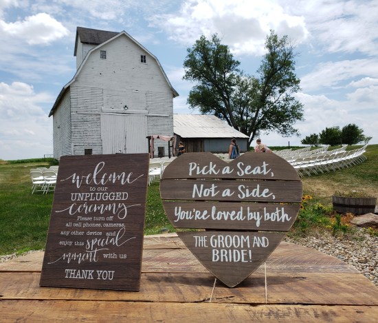 Two wooden signs on an outdoor wooden table. Cutesy writing, folksy language -- one asks us to put away all the devices during the ceremony, the other says we can sit on either side, wherever's cool.