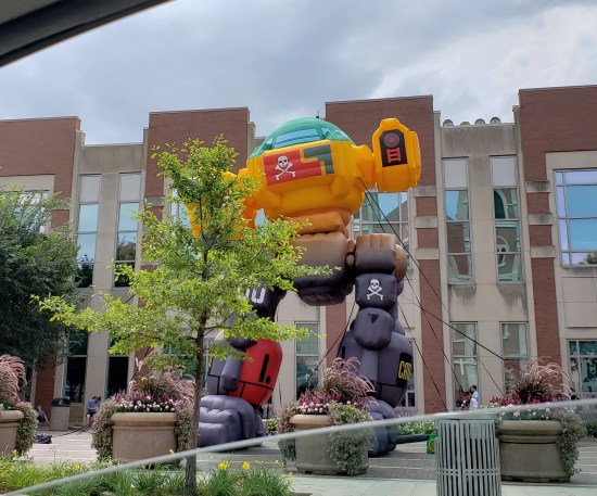 Nine-foot yellow robot statue outside a convention center with trees and benches and other amenities.