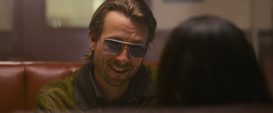 Glen Powell smiling in sunglasses, sitting in a diner.