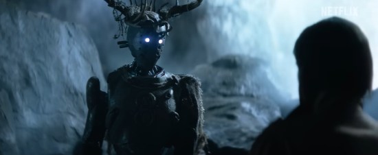 Robot in darkness wearing headgear made of antlers, talking to someone in the shadows.