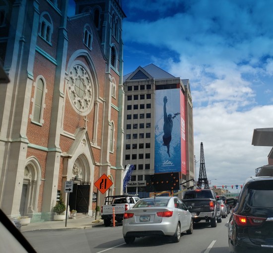 Busy downtown street with a cathedral, a small skyscraper with a big photo of a Black swimmer, and a tiny Eiffel Tower down the street.