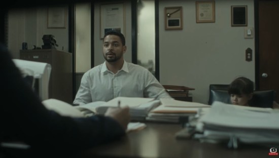 Nervous Latino man in a dress shirt sits on the interviewee side of a desk with paper stacks on it. His tiny daughter's head is barely visible in the next chair over.