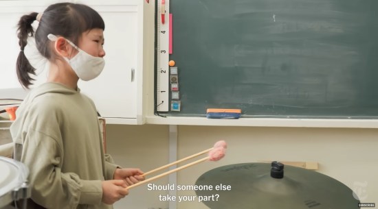 Tiny masked Japanese girl stands over a high-hat cymbal with two mallets with pink fuzzy heads. Teacher says offscreen in subtitles, "Should someone else take your part?"