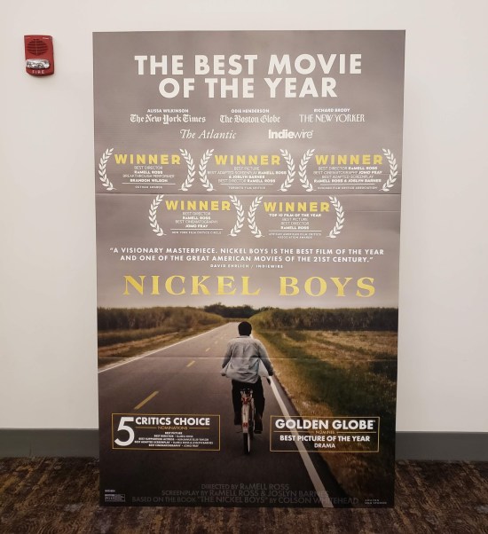 Large standee for the film next to a white theater wall. The image is first-person viewpoint from a kid on a bicycle, riding behind another one, both heading down a straight country road surrounded by fields.